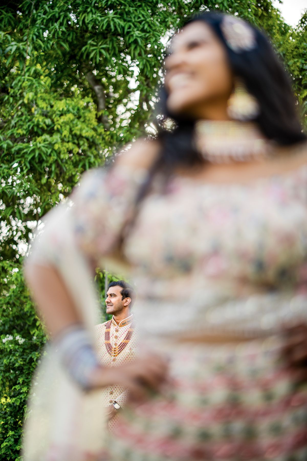 Indian bride in embroidered lehenga smiles outdoors as groom stands in soft focus background during wedding.