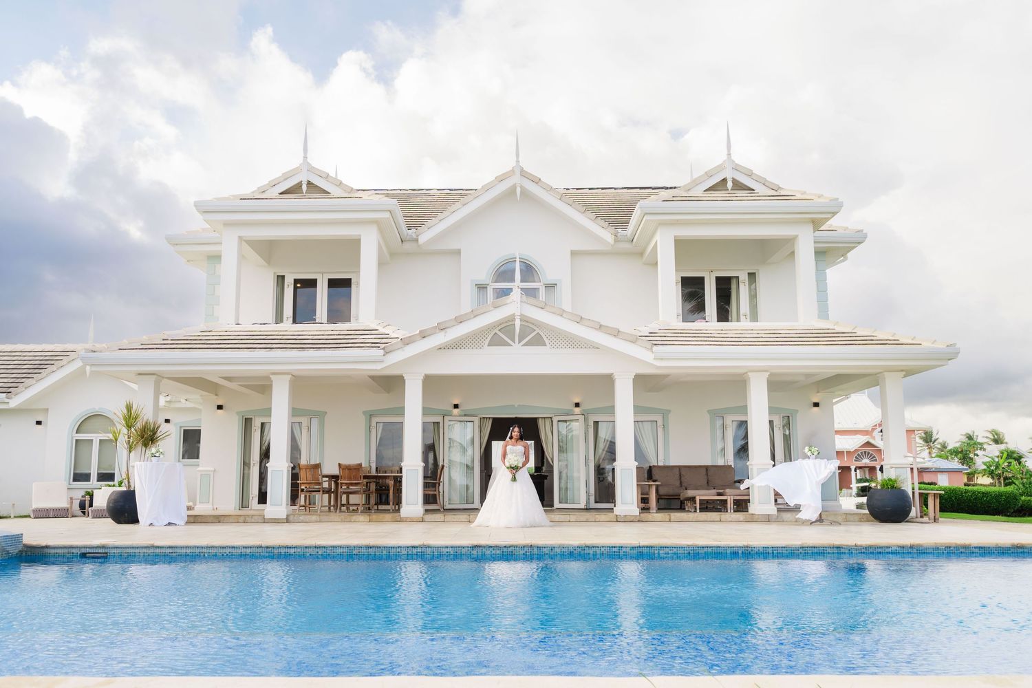 Bride and groom pose by a stunning white luxury villa with a sparkling pool on their wedding day.