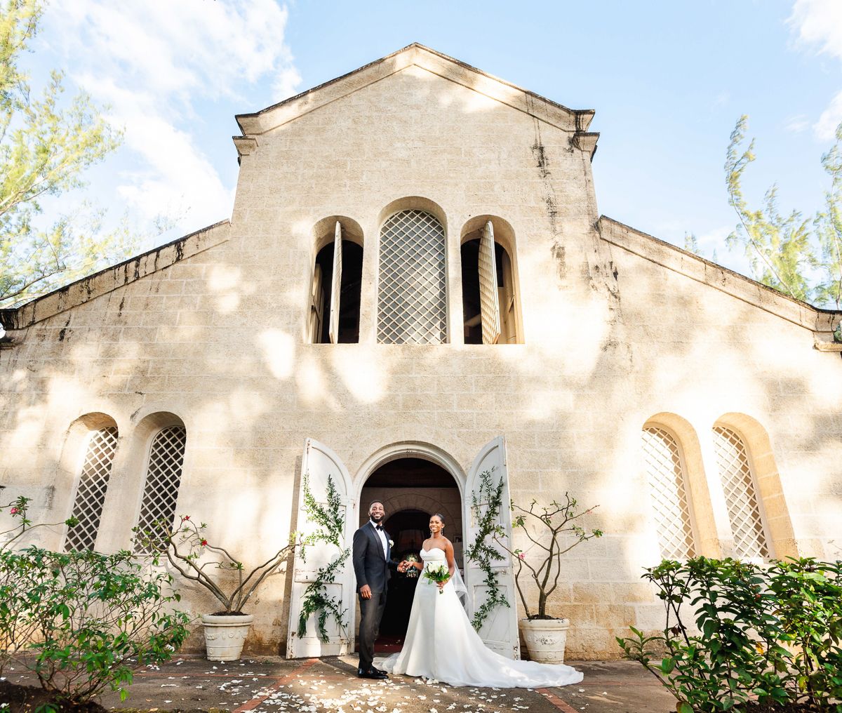 A couple stands at the entrance of a beautiful Mediterranean-style stone church with arched windows and lush greenery, in St James, Barbados