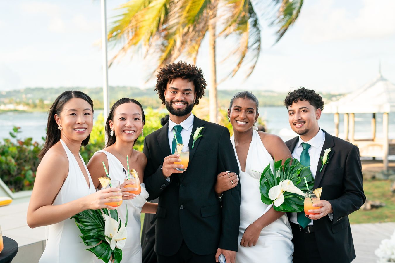 Wedding party joyously celebrates outdoors with drinks and laughter at a tropical destination venue.