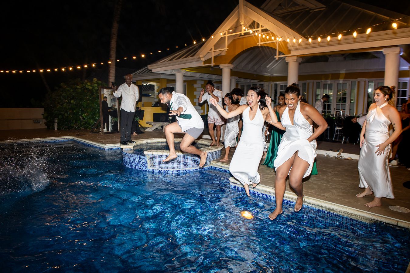 Wedding guests leaping into illuminated swimming pool during nighttime celebration.