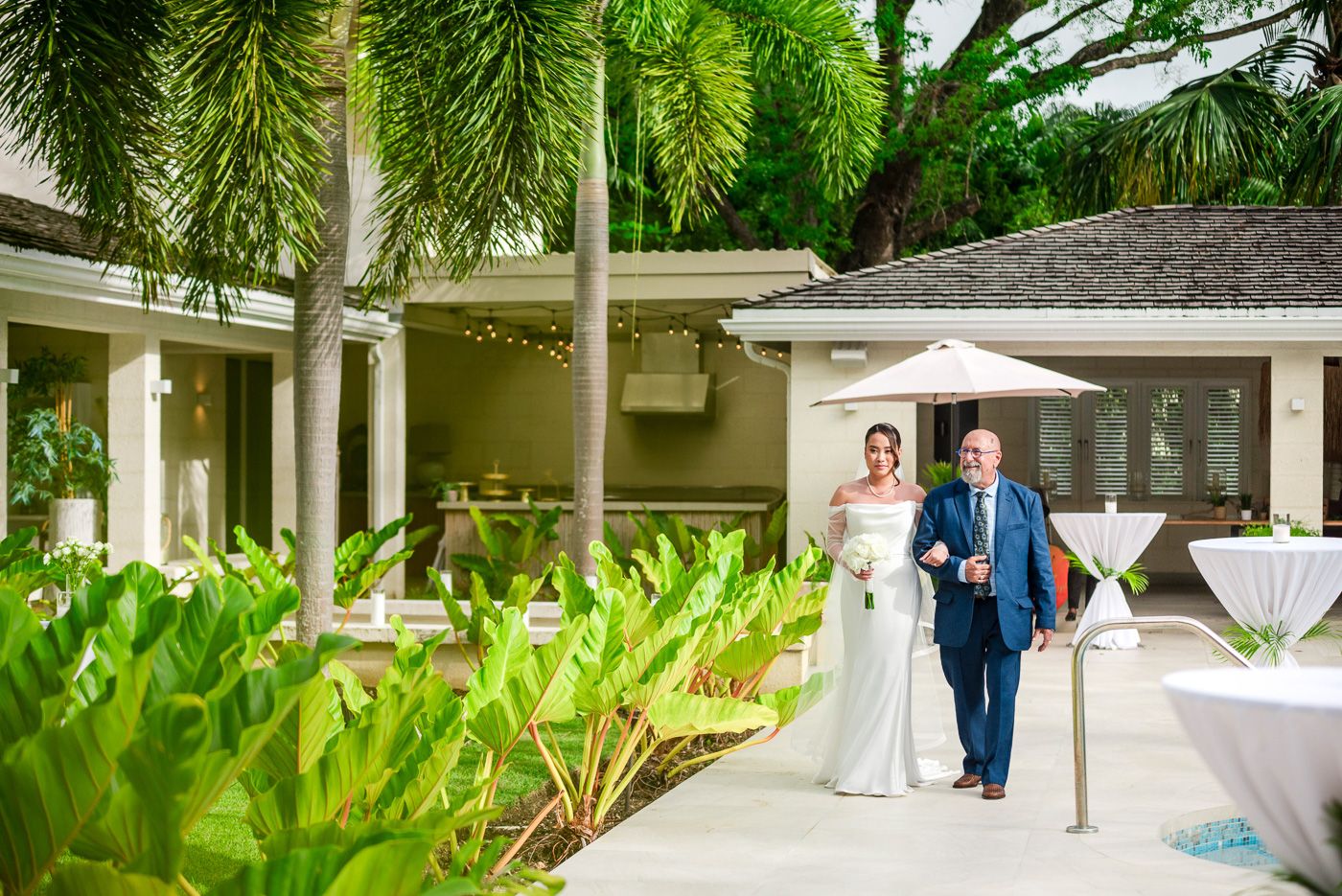 Wedding guests socialize on a white pathway surrounded by lush tropical landscaping.