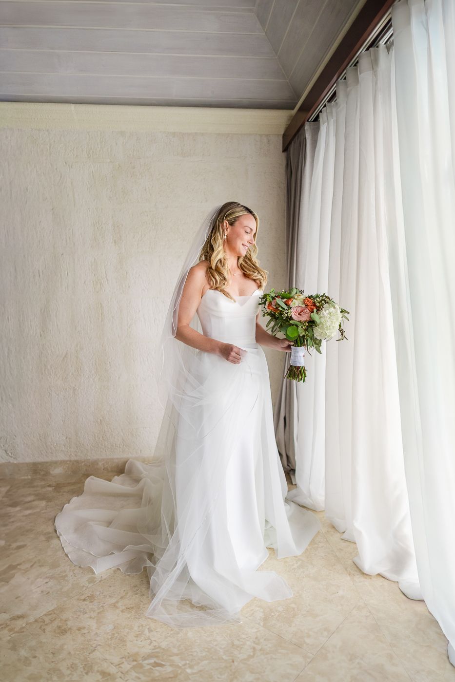 Bride in white gown holding floral bouquet standing by sheer curtains in elegant wedding venue.