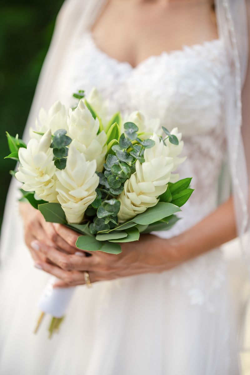 A delicate white peony bouquet with eucalyptus leaves is held against a white wedding dress in natural lighting.