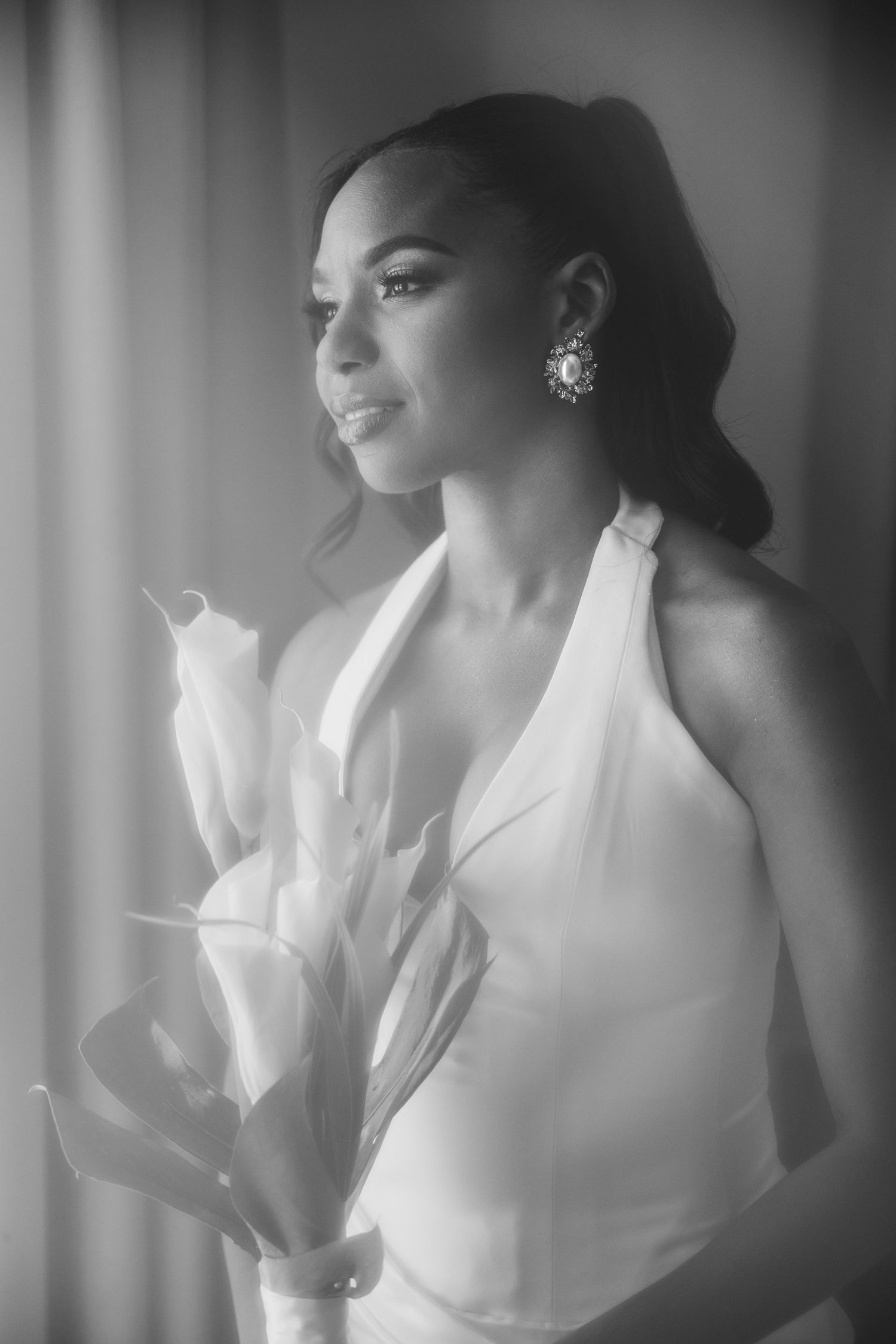 Black and white photo of an elegant bride holding calla lilies, wearing a halter dress and pearl earrings.