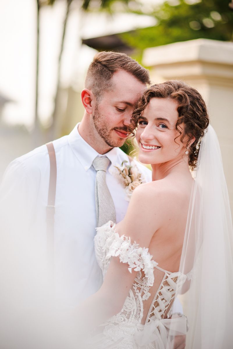 A bride in a lace gown and veil smiles radiantly as the groom in a white shirt and gray tie nuzzles her cheek.