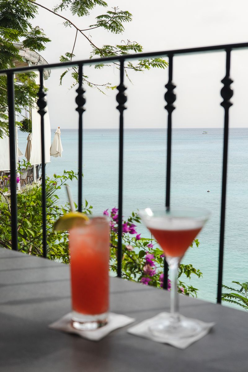 Two tropical cocktails on a balcony table overlooking a turquoise Caribbean sea, surrounded by lush greenery and pink flowers.