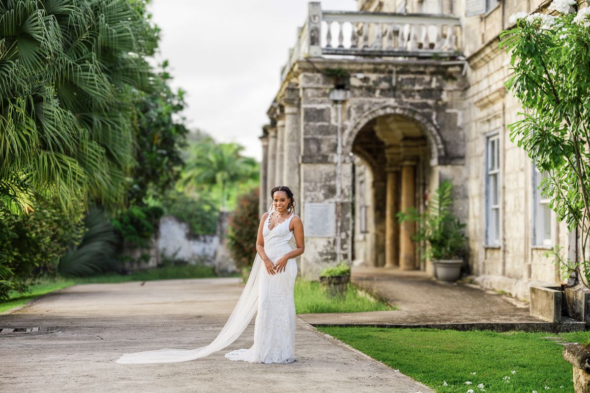 A bride in a white wedding dress poses in front of a stone archway surrounded by lush greenery and gardens.