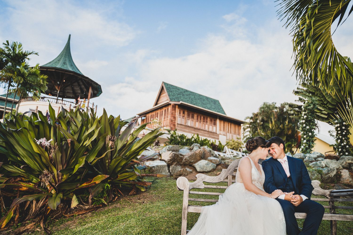 A couple shares a romantic moment in a tropical garden setting with palm trees and pagoda architecture in the background.