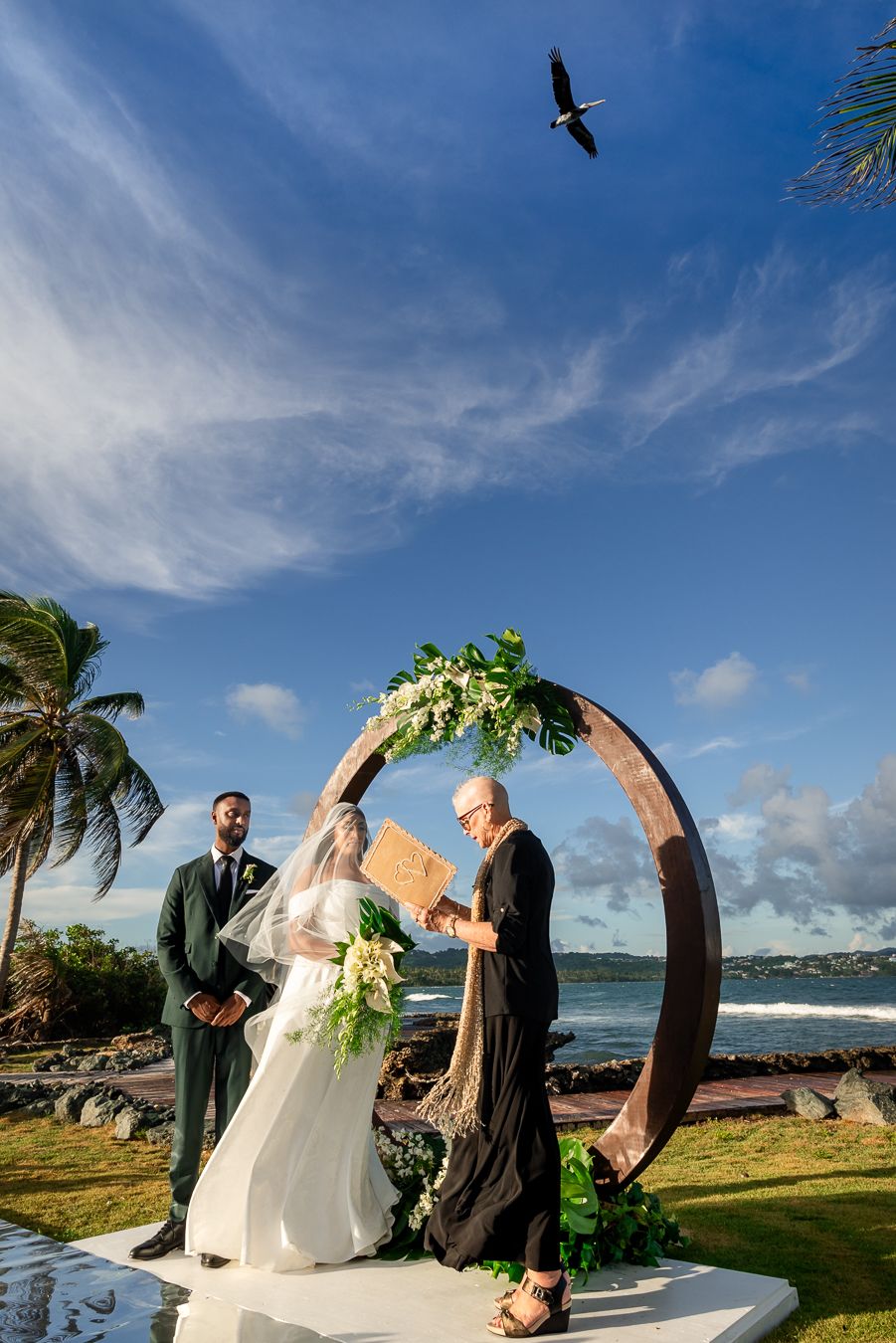 Wedding ceremony under circular arch with bird soaring overhead at oceanfront tropical location.