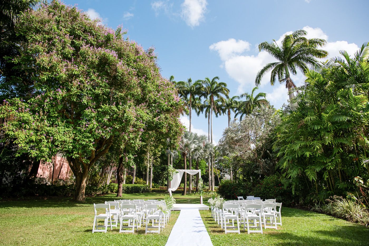 Outdoor wedding ceremony setup with white chairs and palm trees against blue sky.