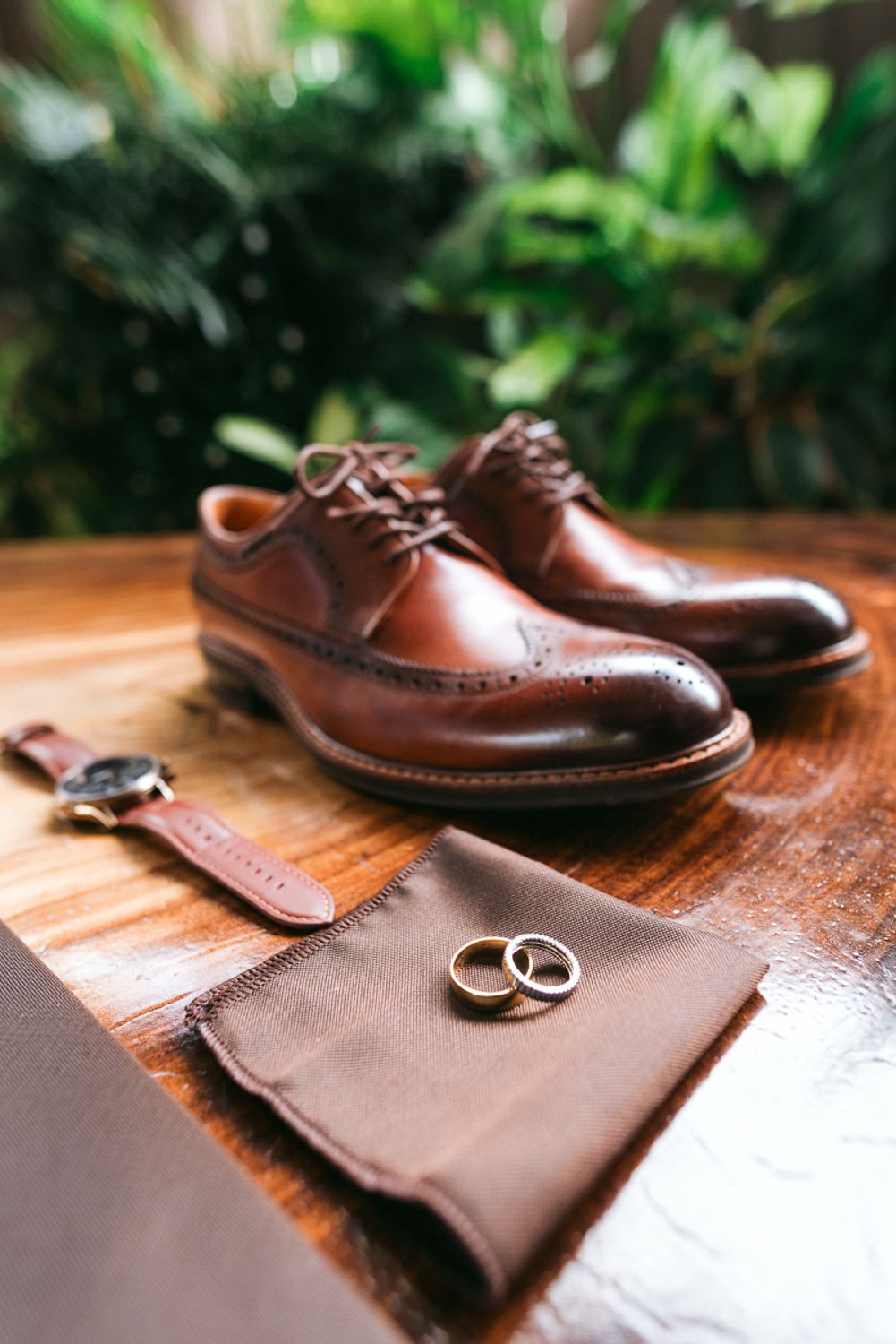 A brown leather dress shoe and silver wedding rings arranged on a wooden surface with a leather wallet in natural lighting.