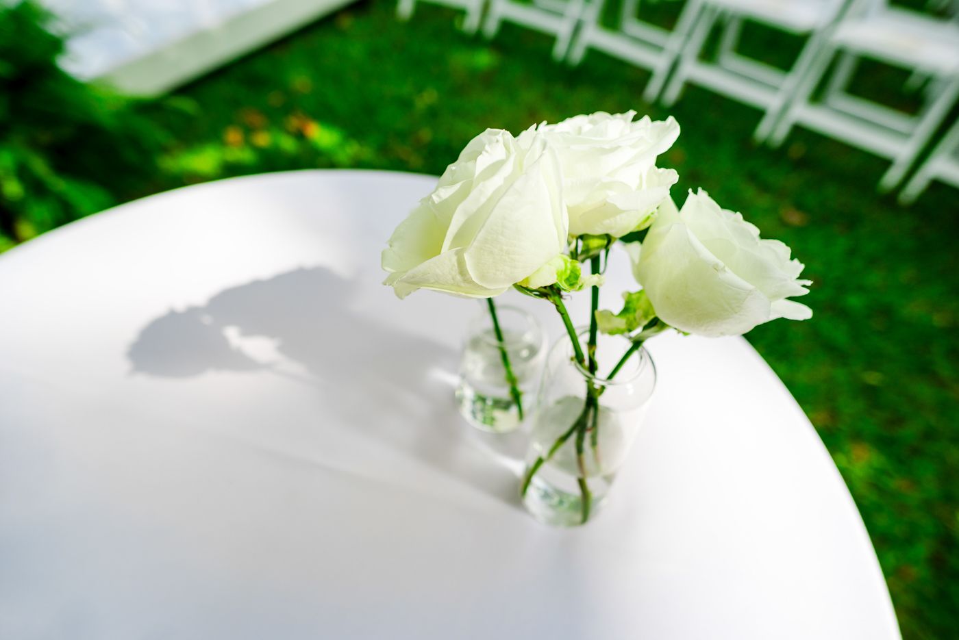 White floral arrangements decorate an elegant outdoor wedding ceremony setup with white chairs in a garden setting.