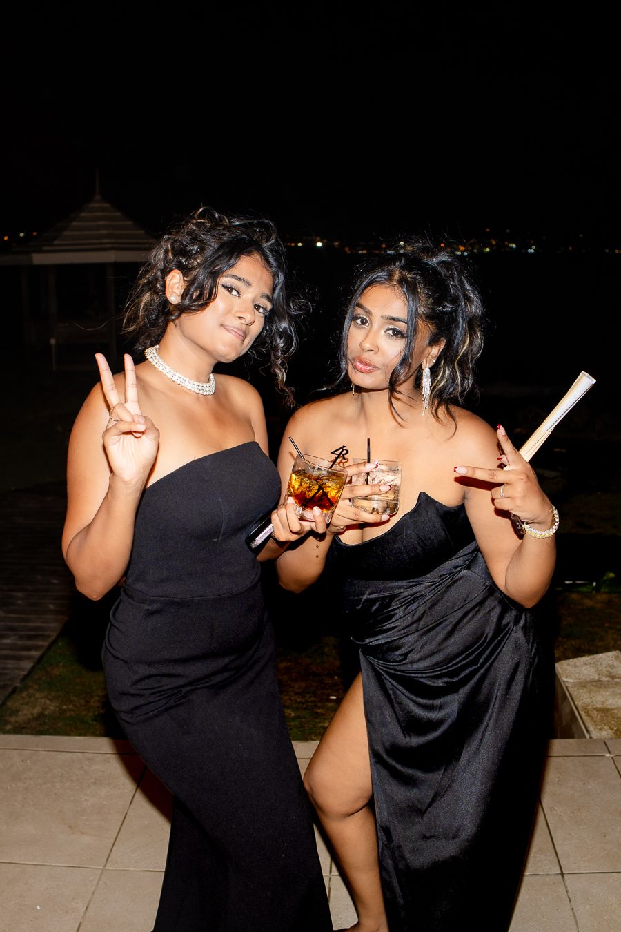 Two guests in black dresses pose with drinks during nighttime celebration.