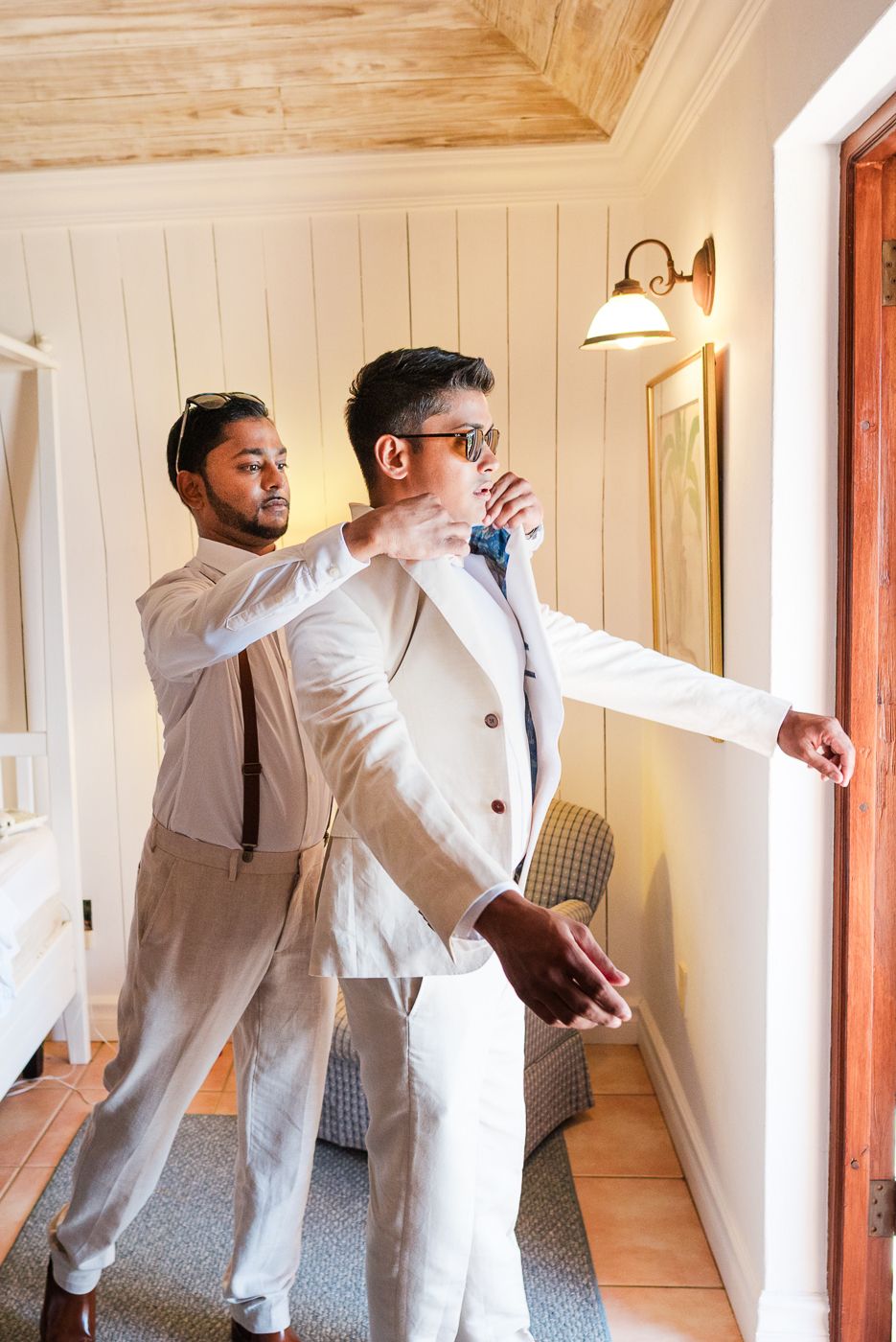 Two people in white suits helping each other get dressed near a window.