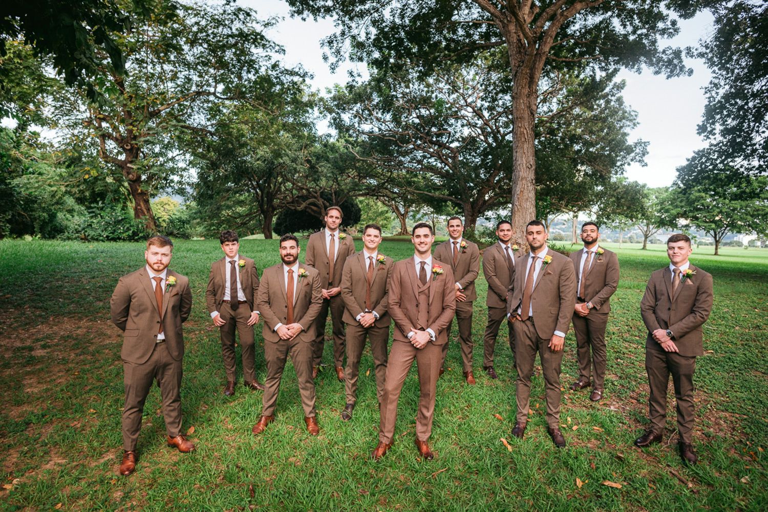 A group of groomsmen in matching brown suits stand together on green grass under tall trees at an outdoor wedding.