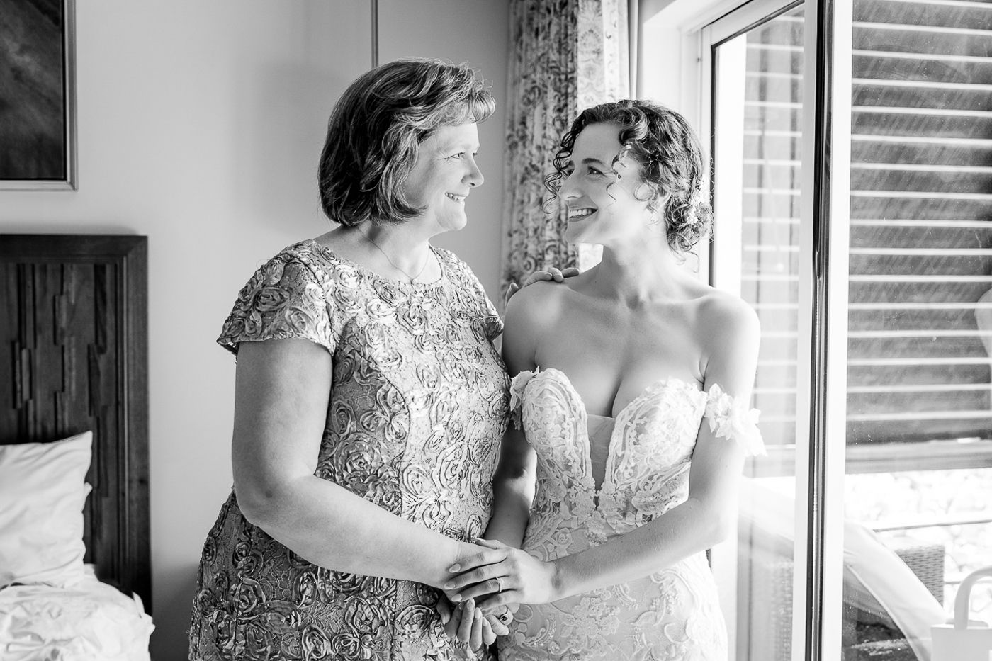 Black and white photo capturing an intimate moment between two formally dressed women by a window.