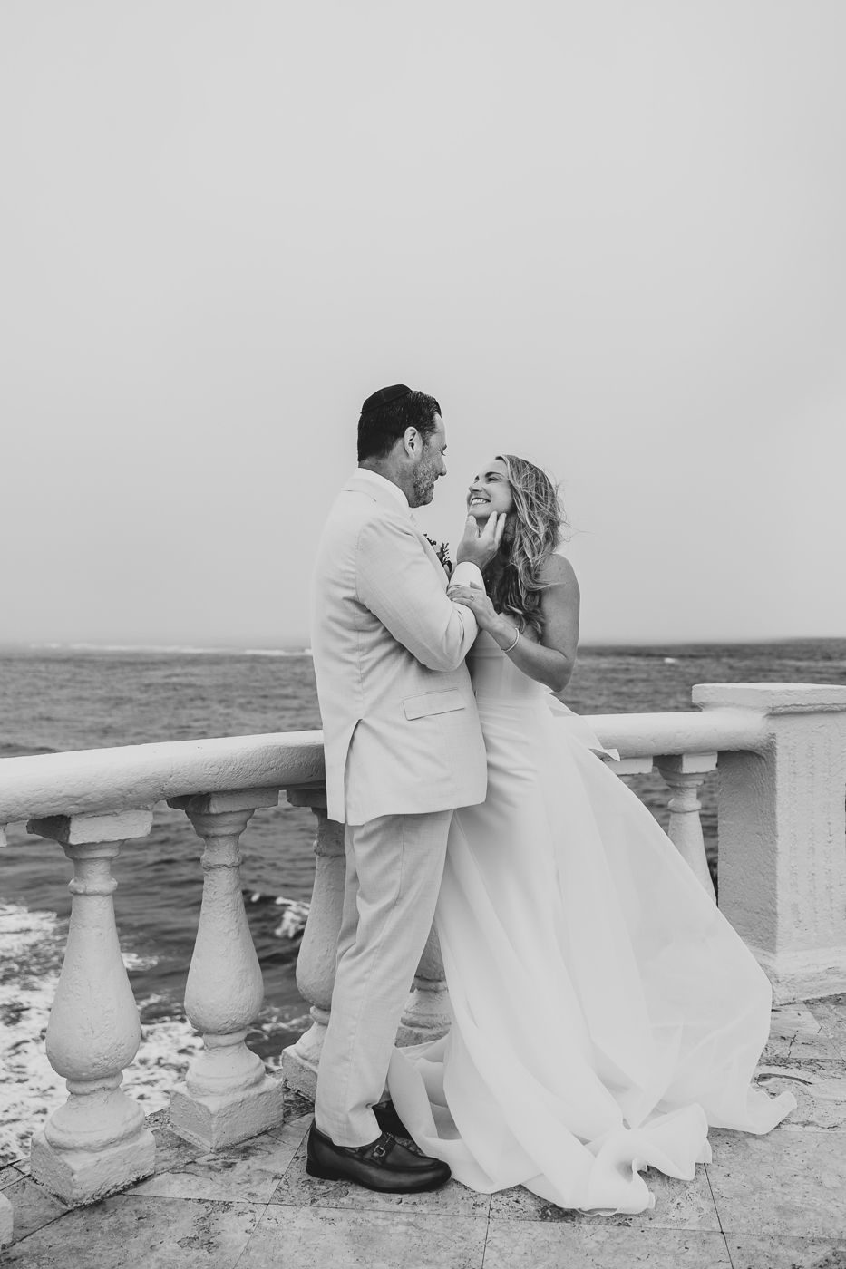 Black and white photo of a bride and groom embracing on a seaside terrace, laughing joyfully on their wedding day.