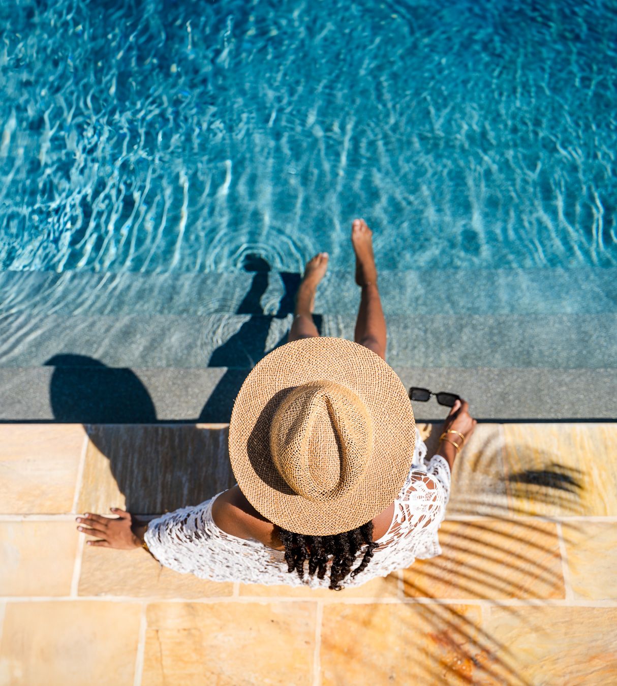 Woman in straw hat sitting poolside with feet in blue water on sunny day.