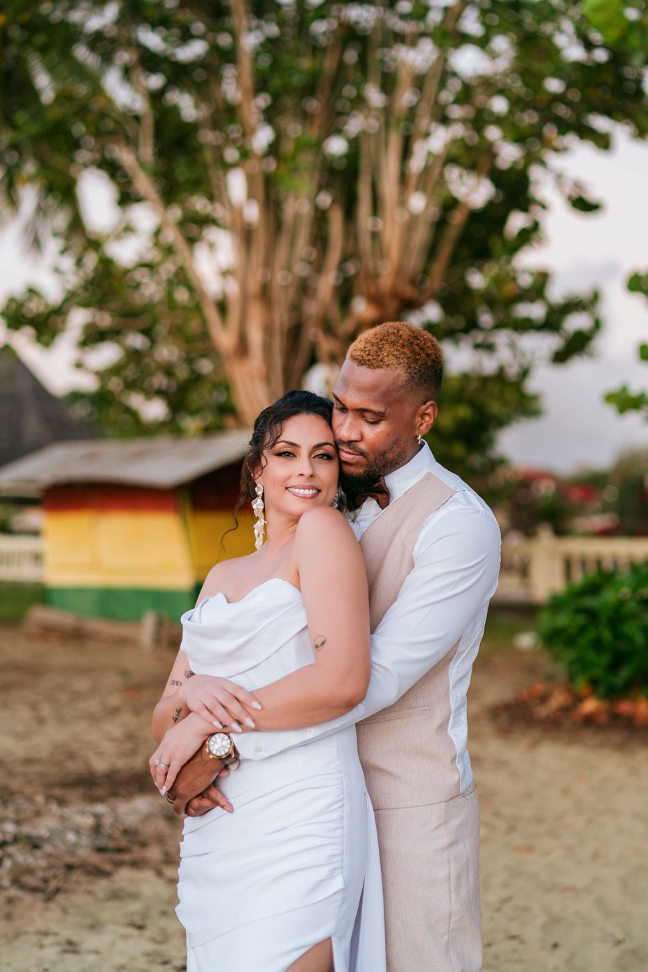 Couple in wedding attire embracing on sandy beach with colorful building and lush tree backdrop.