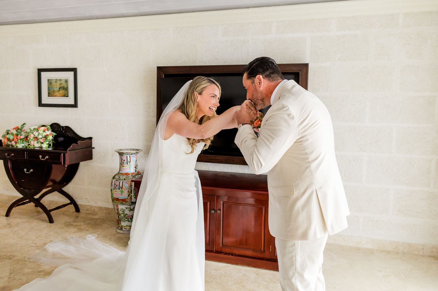 Bride and groom sharing a tender moment indoors, adjusting boutonniere in elegant white wedding attire.