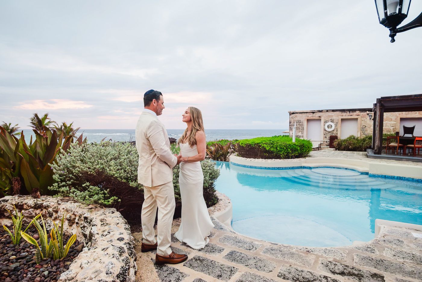 Bride and groom stand by a resort pool with ocean views at sunset in a romantic destination wedding.