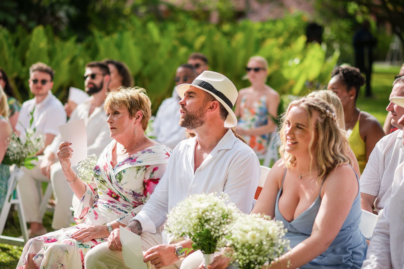 Wedding guests in summer attire seated outdoors watching ceremony in tropical garden setting.