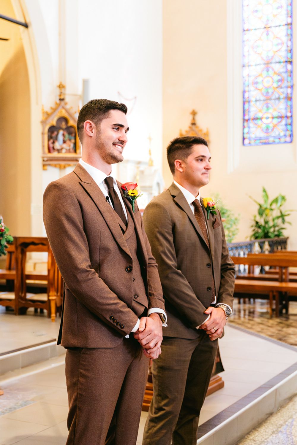Two groomsmen in brown suits stand at attention during a church wedding ceremony.