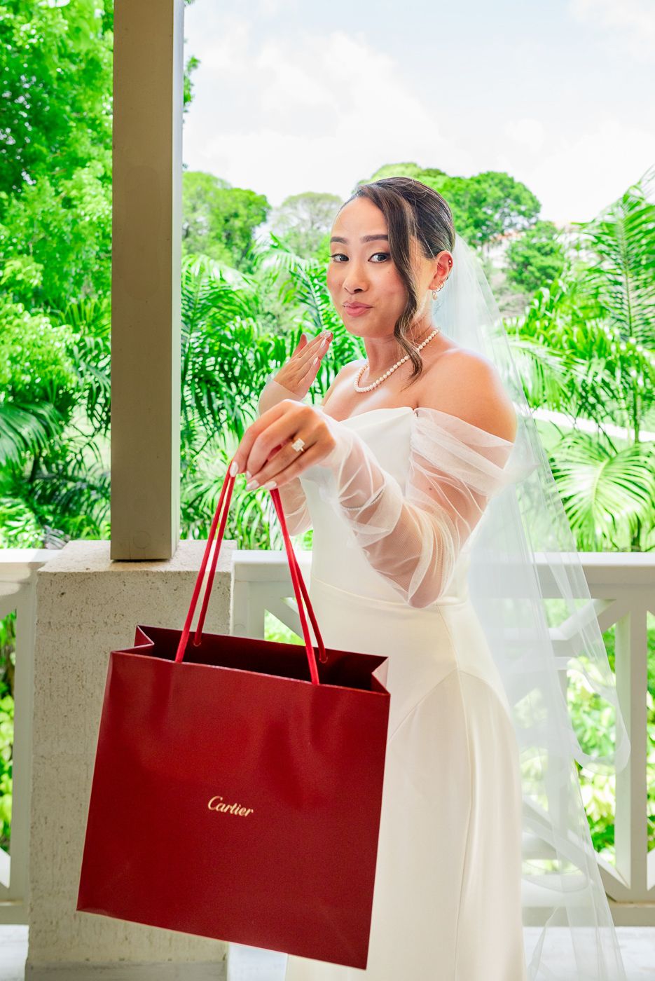 A classic white wedding gown is paired with a red Cartier shopping bag on a sunlit veranda.