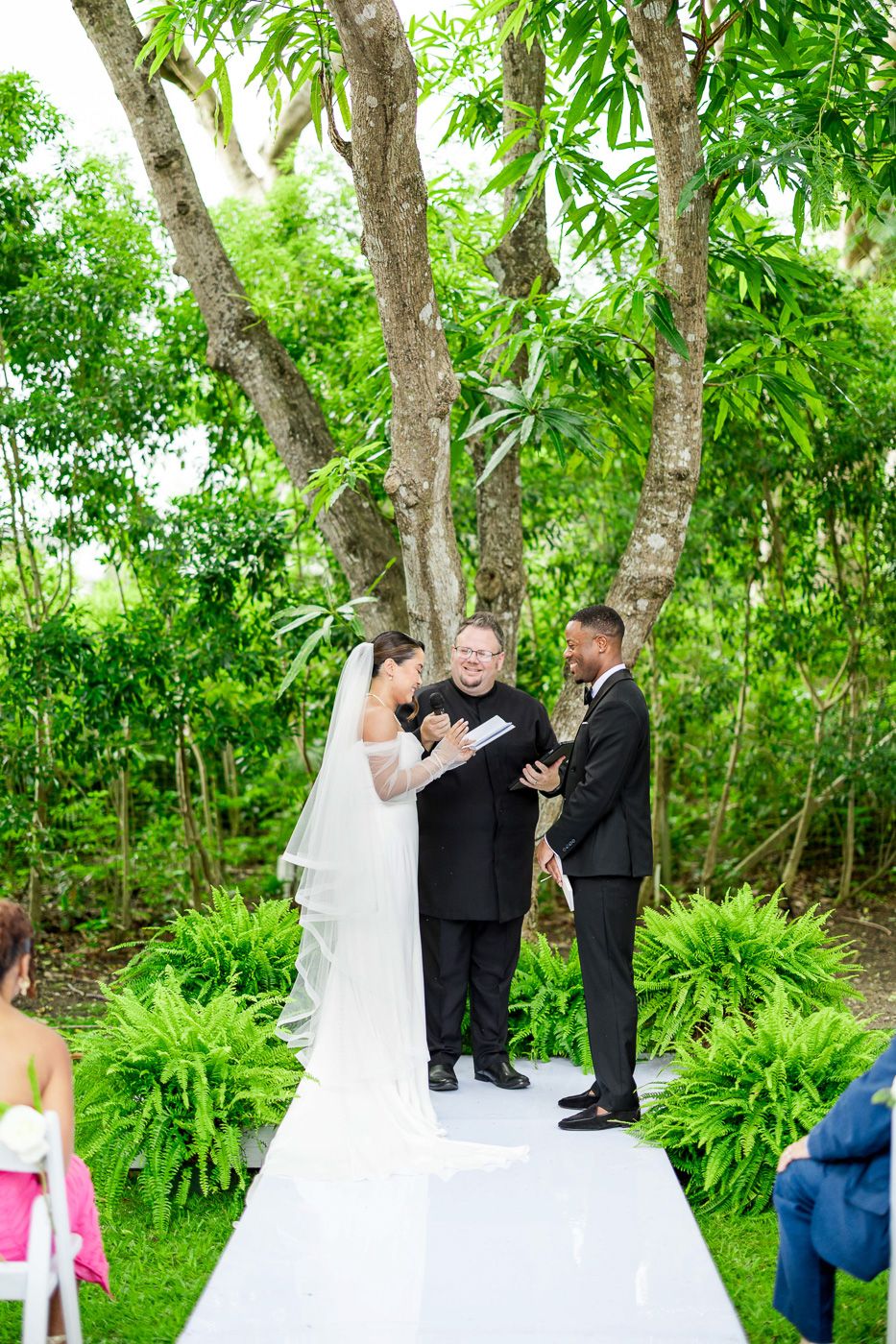 An intimate wedding ceremony takes place on an elevated white platform surrounded by lush tropical foliage.