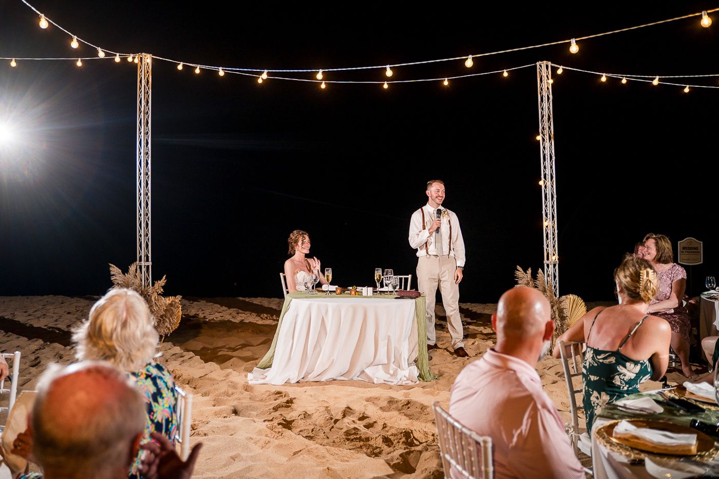 Beach wedding reception with string lights and white-clothed tables along the shoreline.