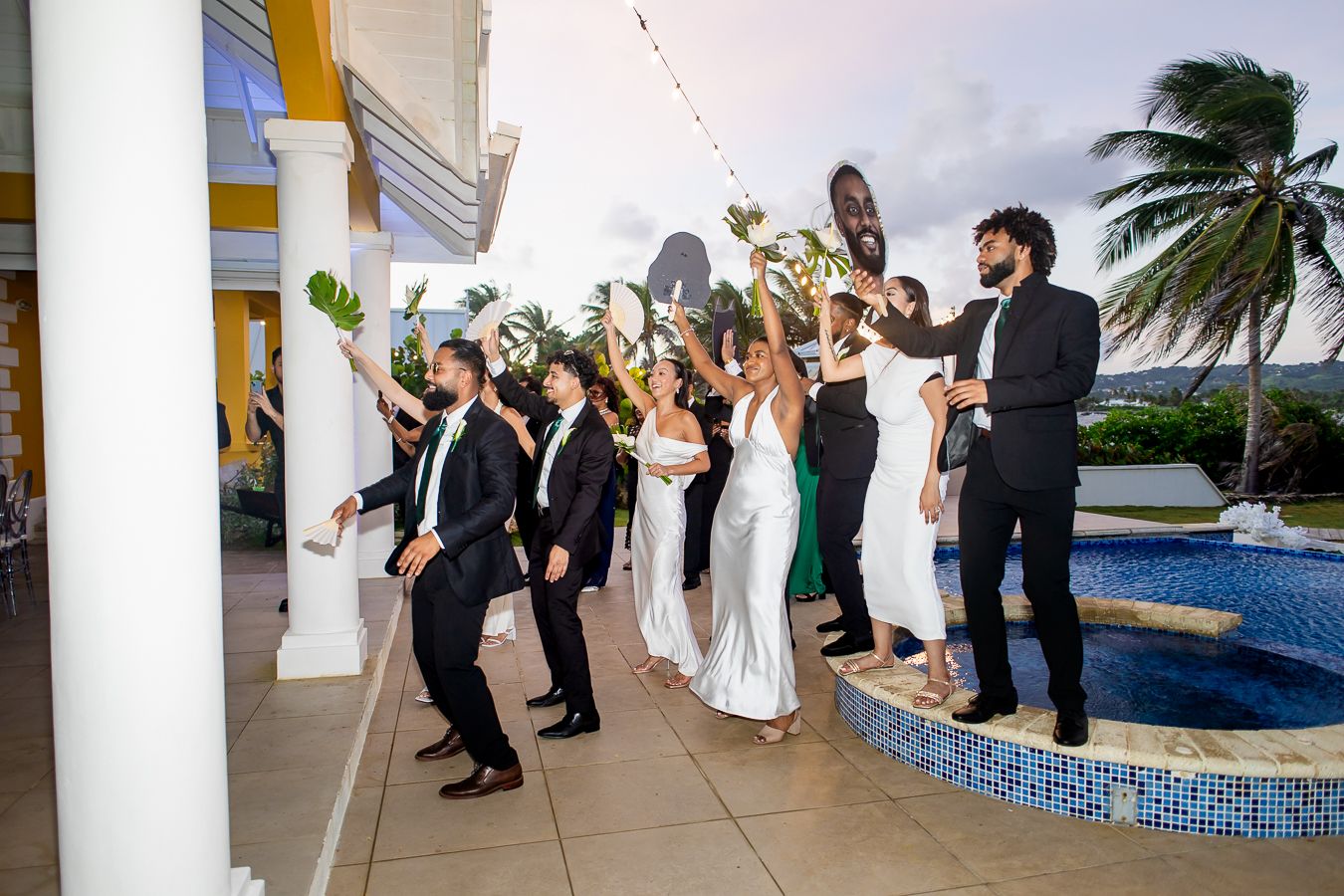 Wedding party dancing beside pool at tropical venue with white columns and palm trees.