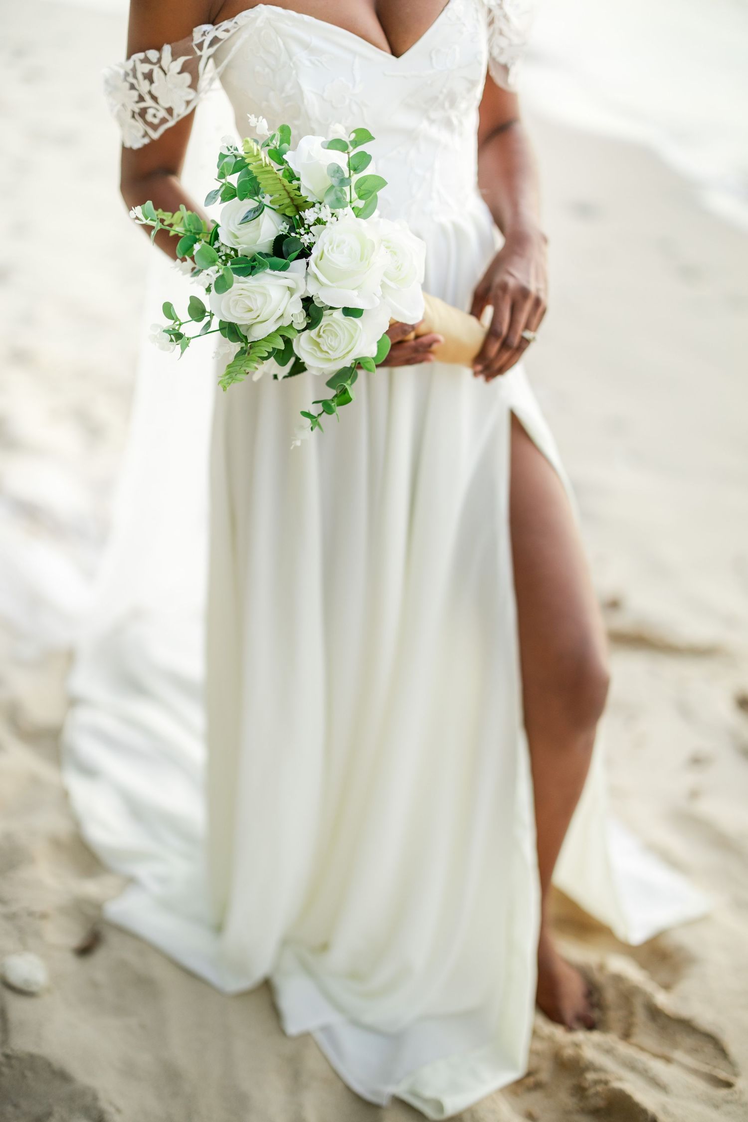 White wedding dress detail with flowy fabric and delicate green and white floral bouquet against sandy beach backdrop.