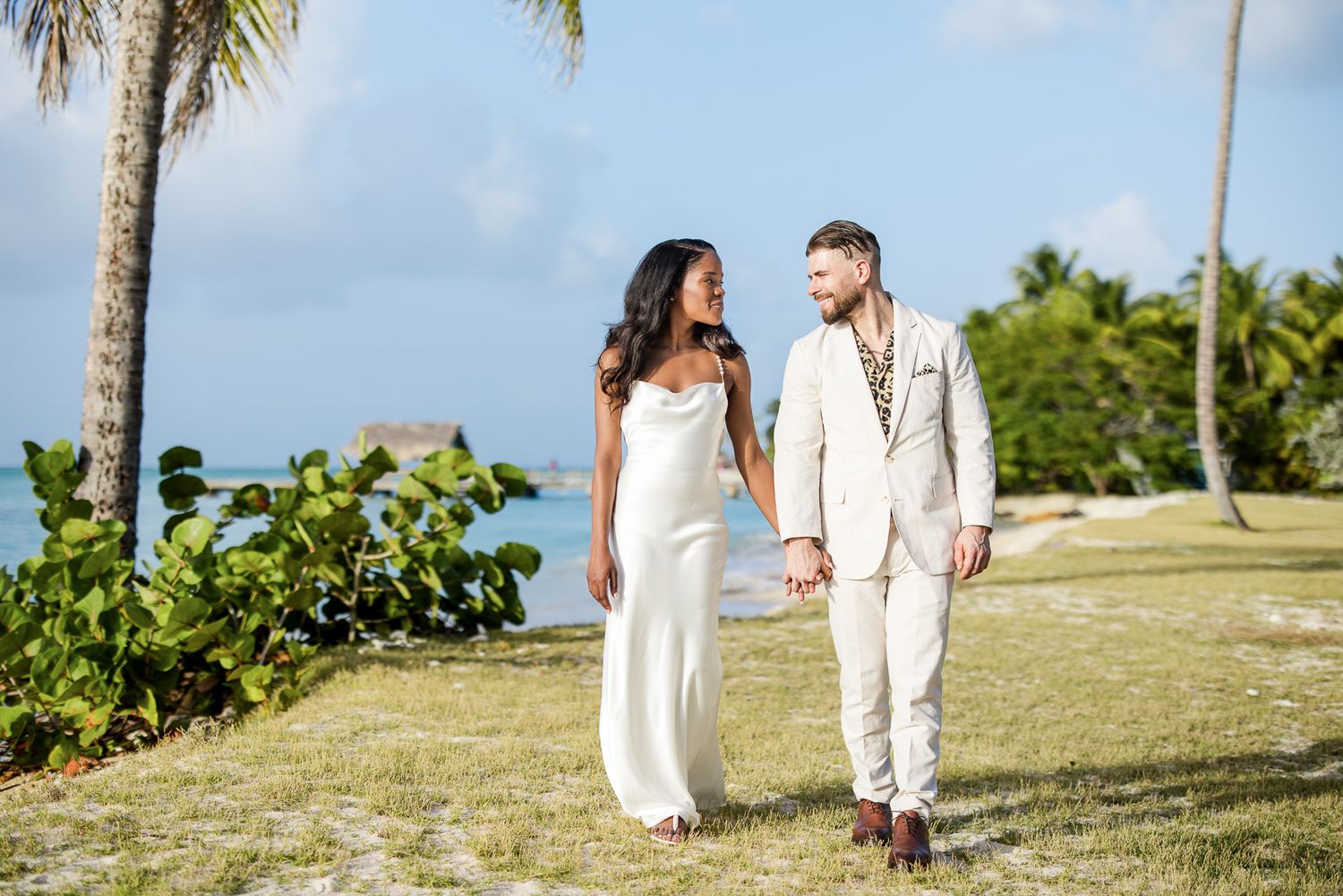 Wedding couple walking along tropical beach with palm trees and blue sky.