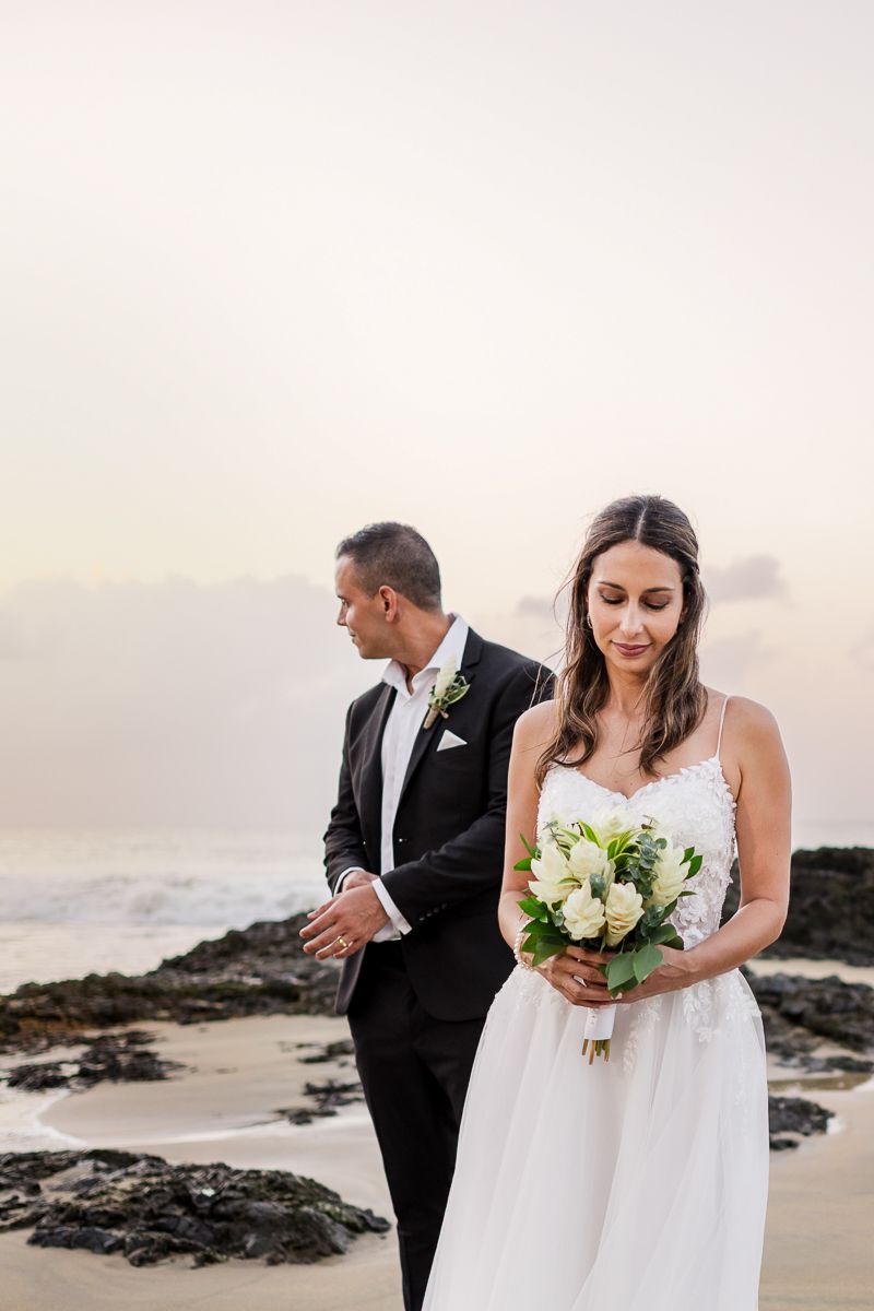 Bride holding white bouquet in wedding dress stands with groom in black tux on a rocky beach at sunset.