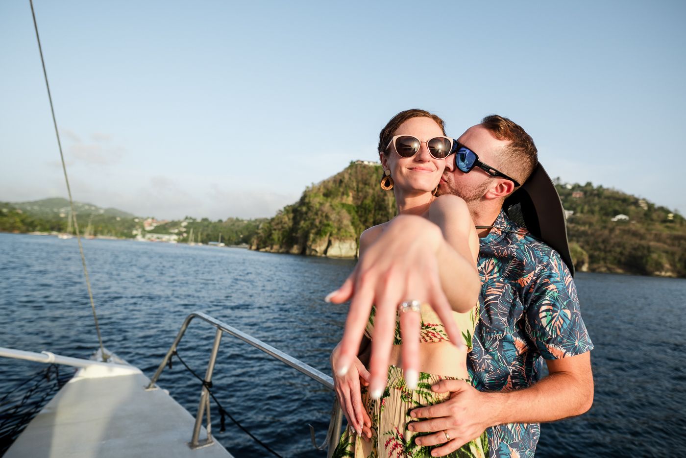 Couple on a yacht showing off engagement ring, hugging and wearing sunglasses with scenic water and hills behind them.