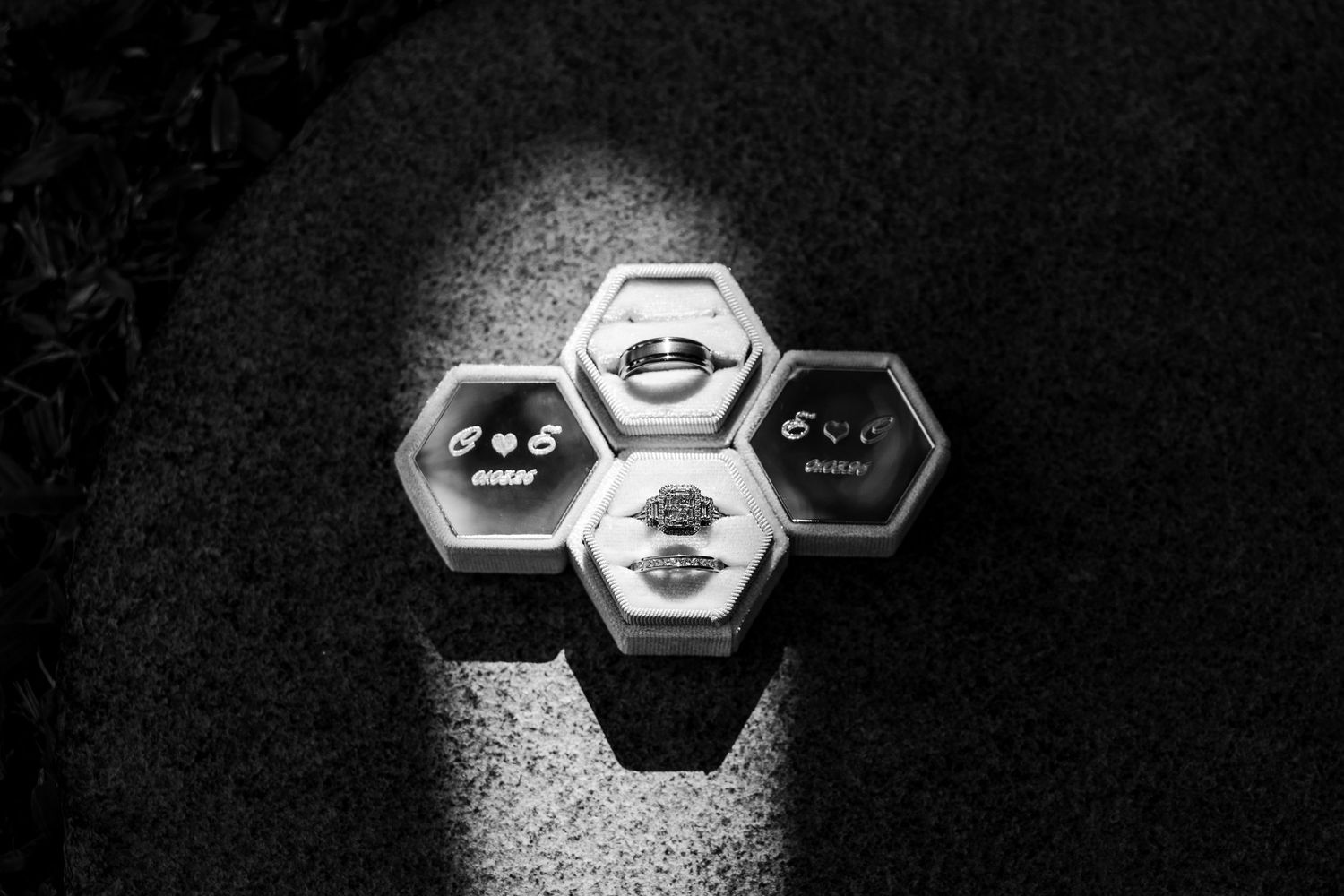Black and white photo of wedding rings displayed in hexagonal velvet boxes on a dark textured surface.