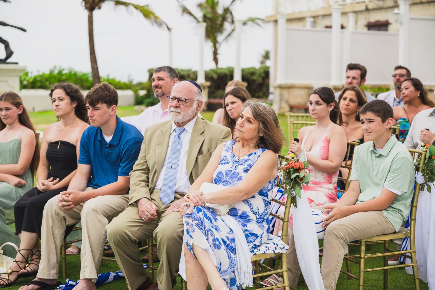 Wedding guests seated outdoors at a beachside ceremony, dressed in elegant attire, enjoying the celebration.