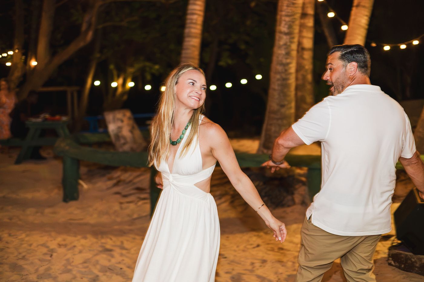 Couple dancing on a sandy beach at night with string lights and palm trees in the background.
