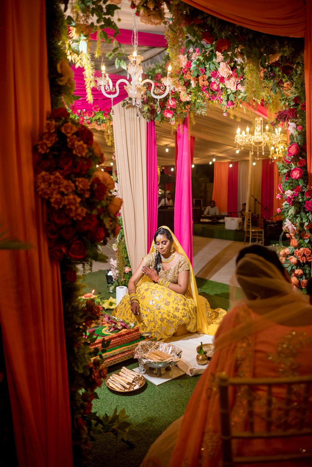 Bride in yellow lehenga sits at floral mehndi ceremony decor with pink drapes, chandeliers, and lush flower arrangements.