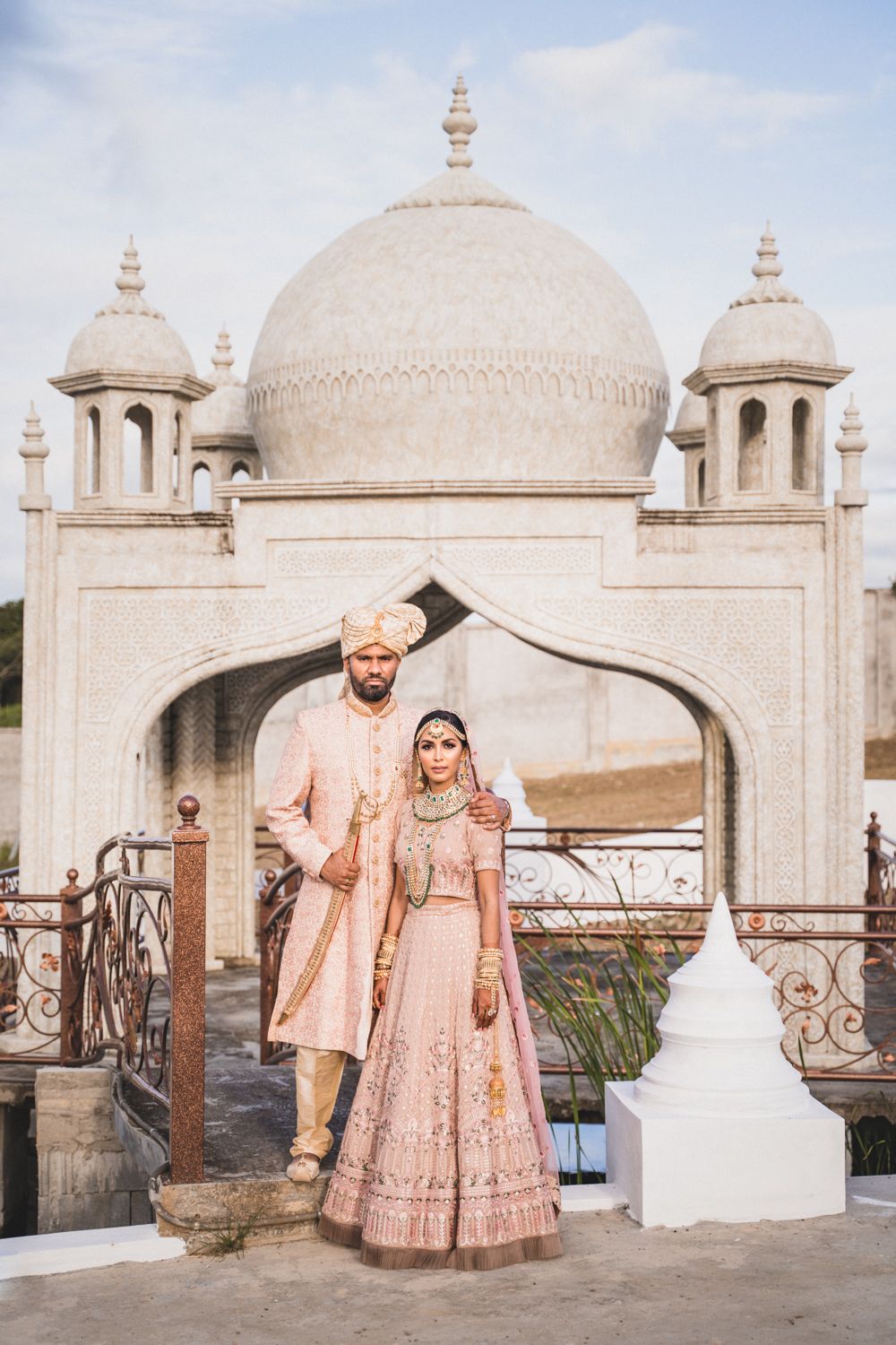 Indian bride and groom in pink wedding attire posing before a white domed Mughal-style arch structure.