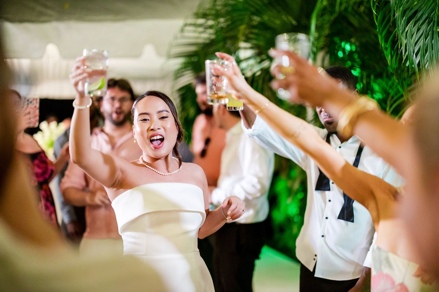 Guests dance energetically at a wedding reception with tropical greenery in the background.