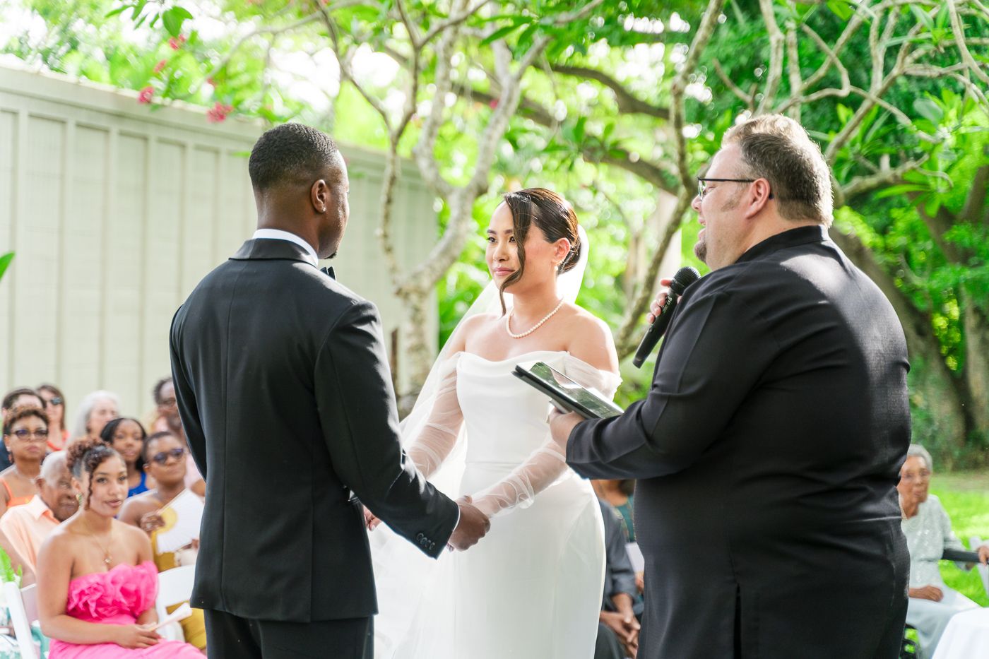 A wedding ceremony takes place outdoors with the couple and officiant standing beneath lush green trees.