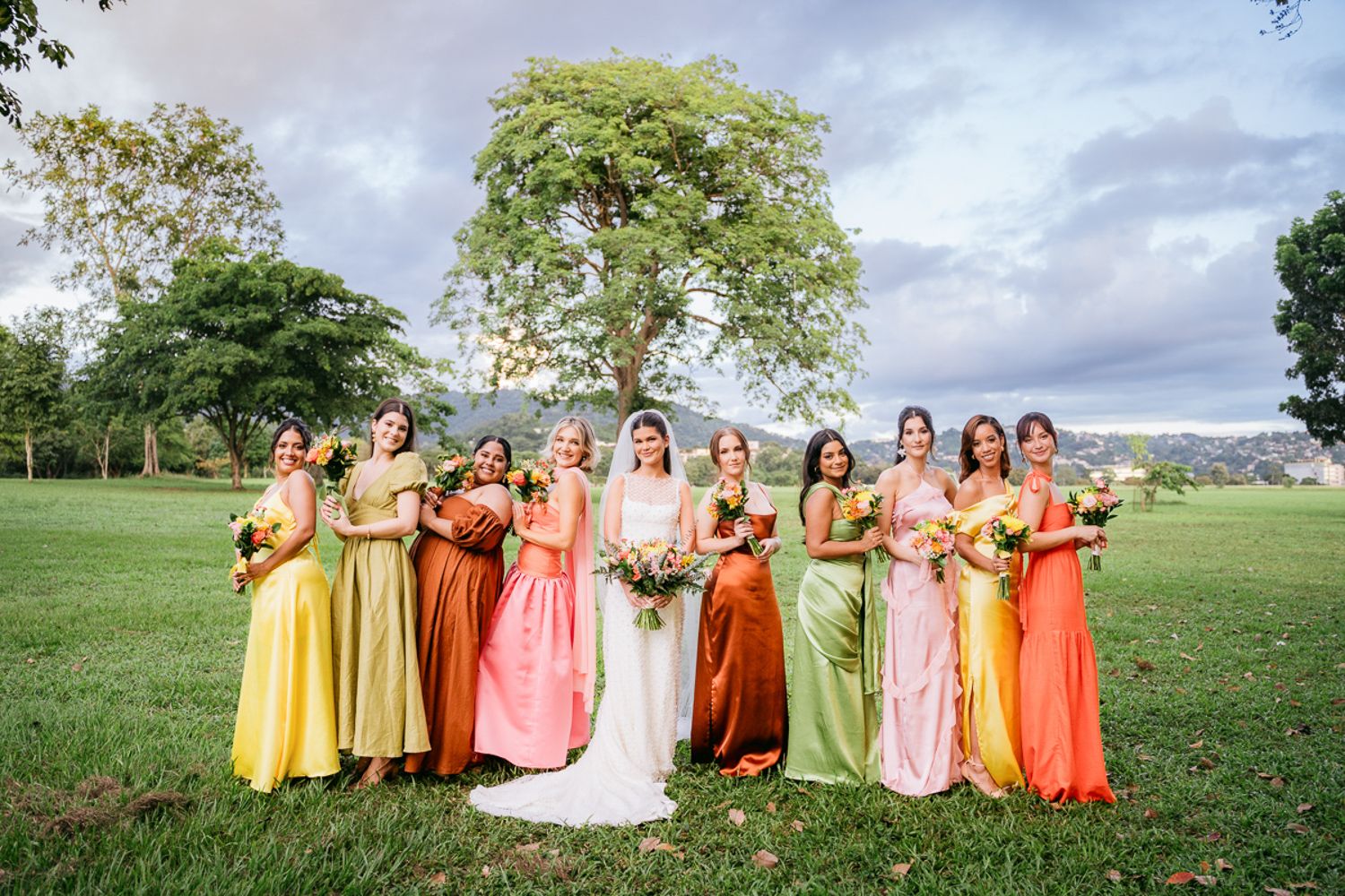 Bridal party in colorful pastel and warm-toned dresses stands together on green lawn under cloudy summer sky.