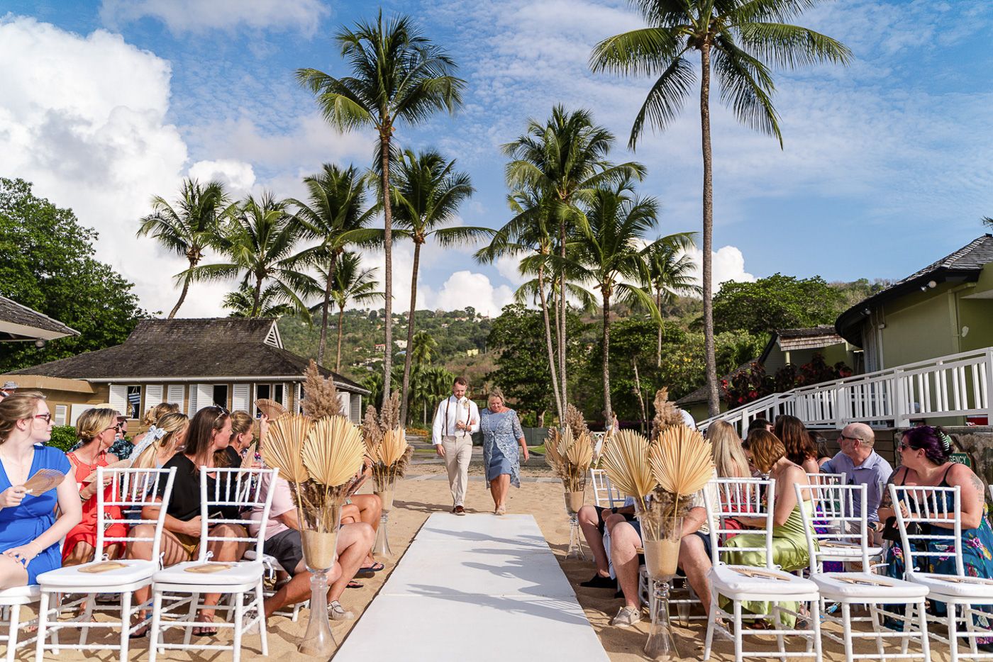 Tropical beach wedding ceremony setup with white chairs and palm trees under blue skies.