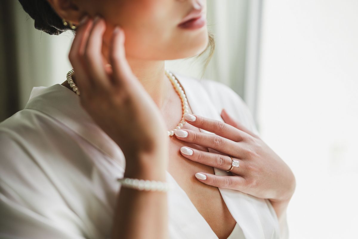 Bride wearing pearl necklace, bracelet, and diamond ring in white robe, touching jewelry elegantly.