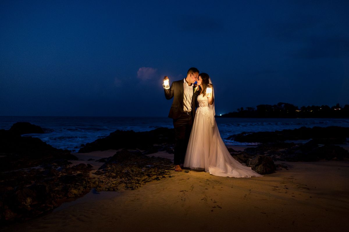 Bride and groom share a romantic kiss on a beach at night, holding torches with ocean waves in the background.