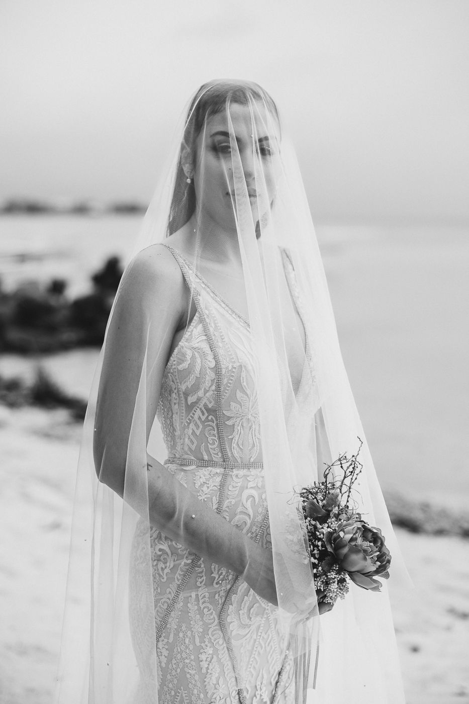 A bride in a lace wedding dress stands on a beach holding a bouquet while wearing a flowing veil in black and white.