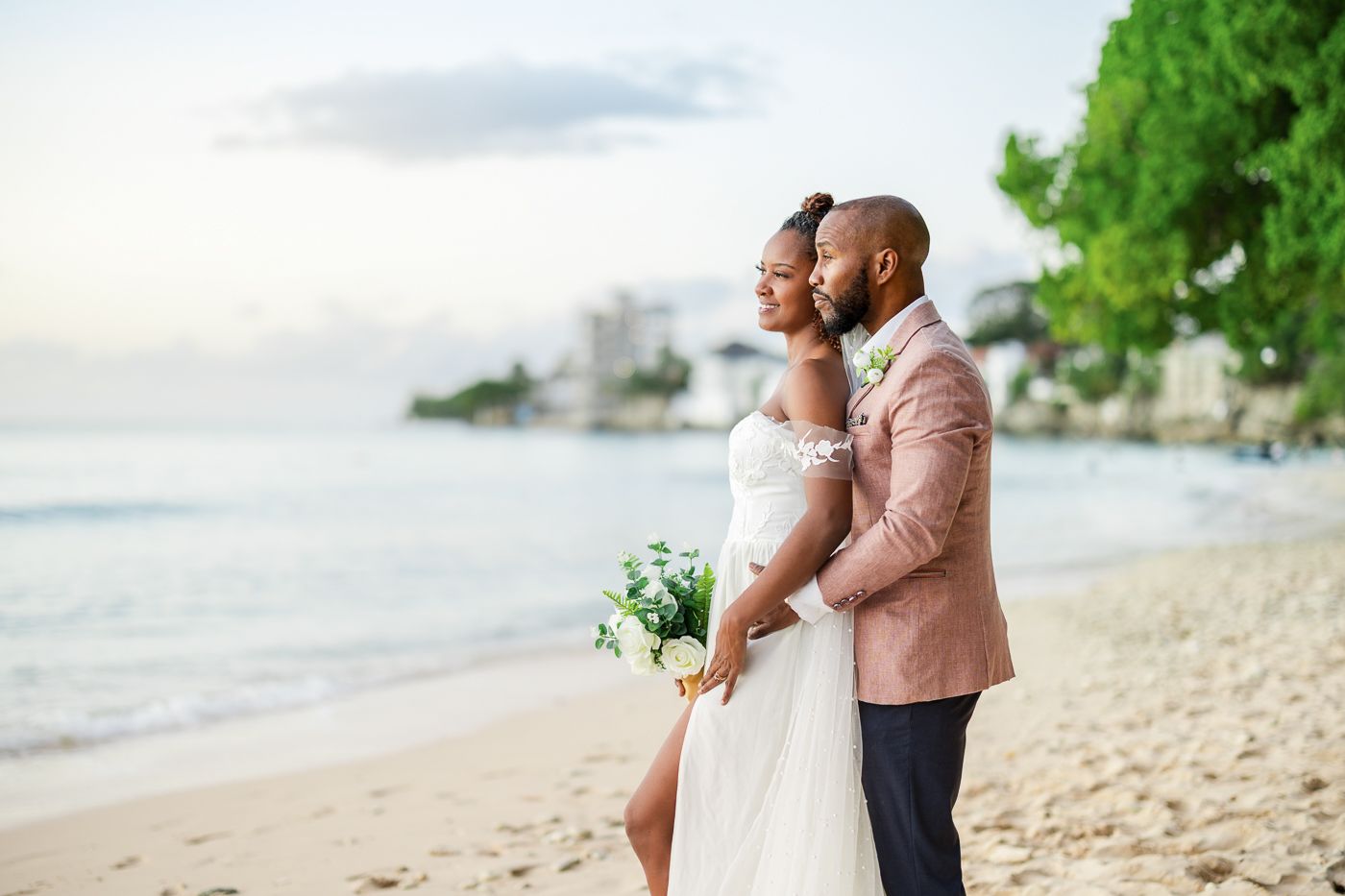 A romantic beachside wedding couple embraces while standing on white sand with ocean waves and greenery in the background.