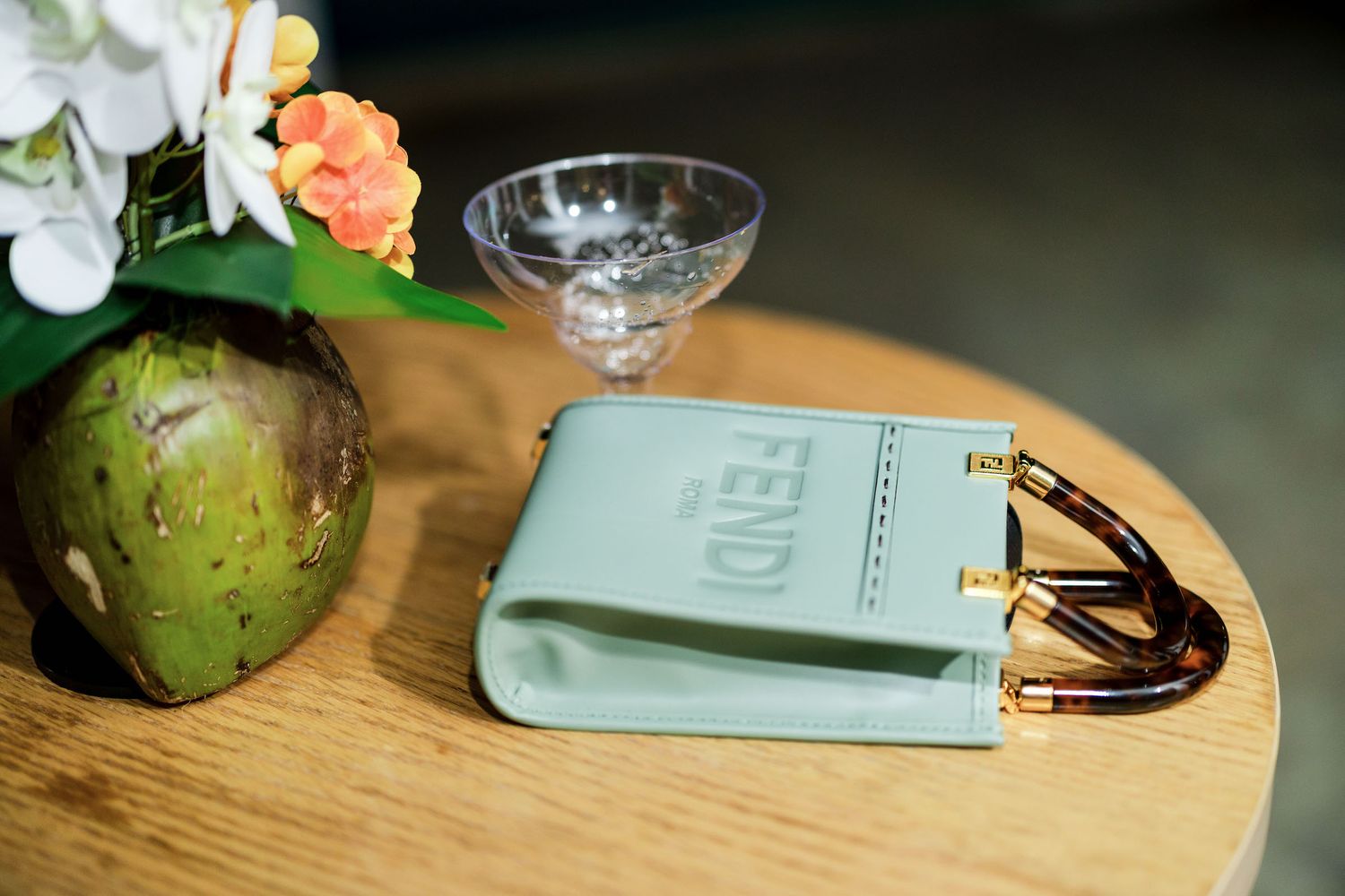 A vintage cocktail glass sits beside a mint green passport holder and coconut on a wooden surface with flowers.